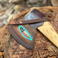 A leather axe sheath with a colorful feather design, shown resting on a rustic tree stump outdoors.