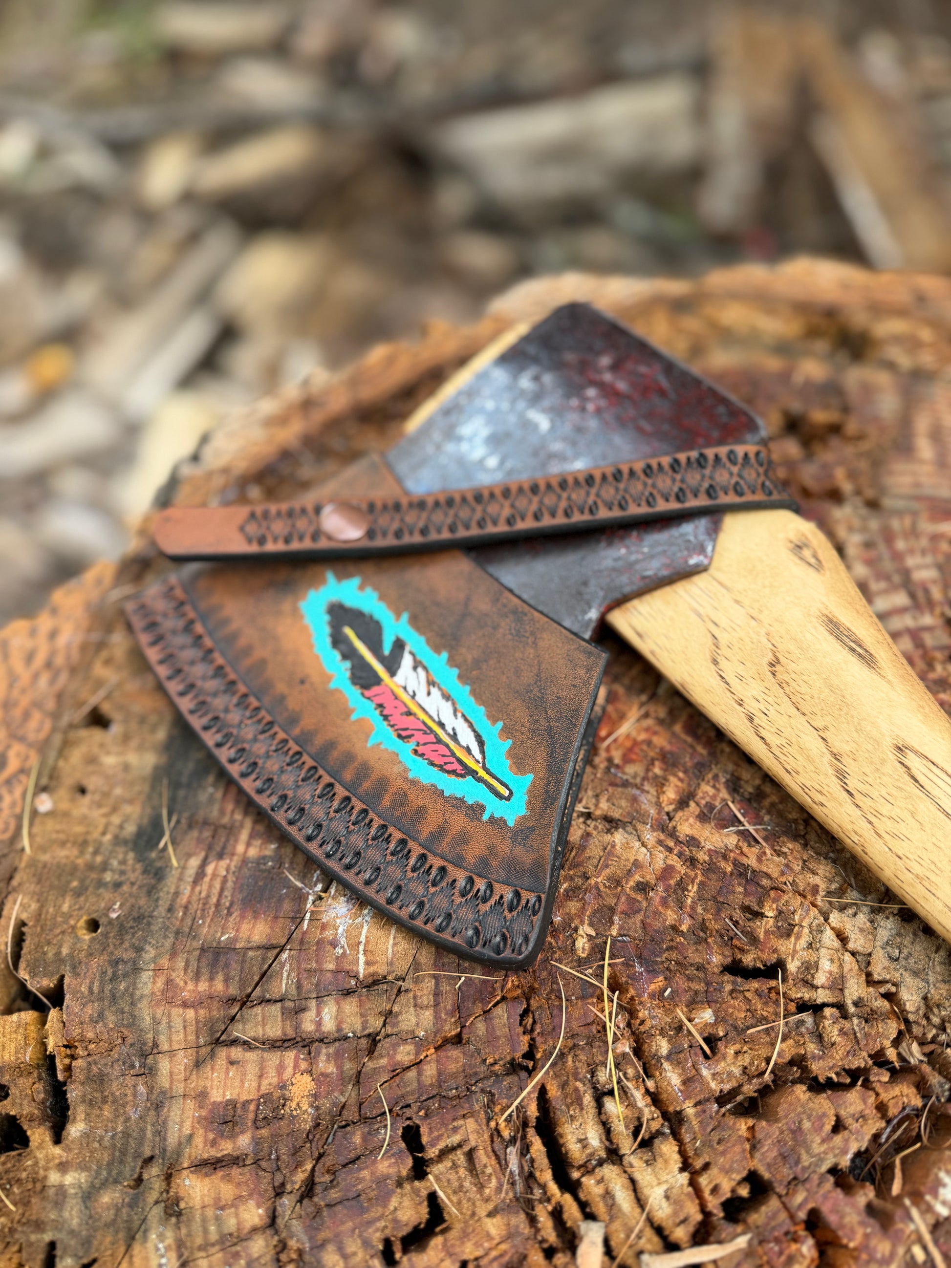 A leather axe sheath with a colorful feather design, shown resting on a rustic tree stump outdoors.