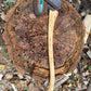 An axe with a hickory wood handle and a dark metal head featuring a turquoise feather accent, resting on a large tree stump surrounded by wood chips and bark.