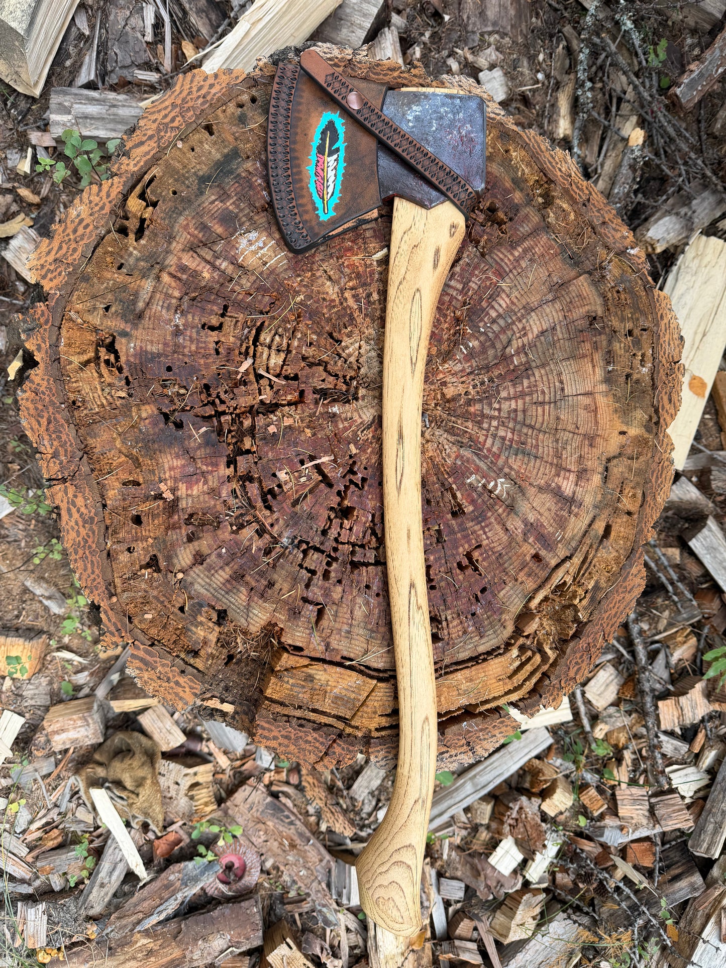 An axe with a hickory wood handle and a dark metal head featuring a turquoise feather accent, resting on a large tree stump surrounded by wood chips and bark.