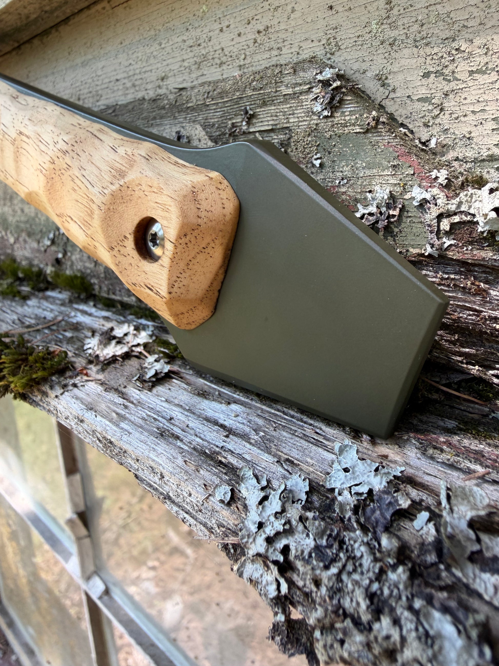 A close-up of a hand axe or hatchet with a natural wood handle and an olive green metal blade, resting against a weathered wooden surface outdoors.